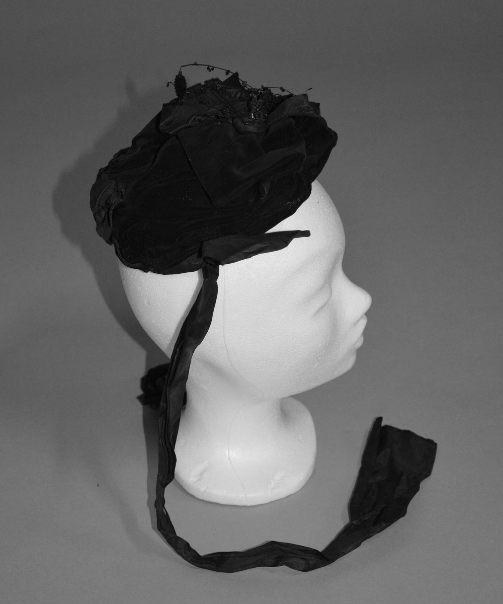 Kysehatt i sort fløyel pyntet med rosett av silkebånd og et motiv med sorte perler og paljetter. To knytebånd i sort silke festet med rosett ved ørene. Opprubbet silkebånd i nakken. Dobbelt fôr i sort bomull.