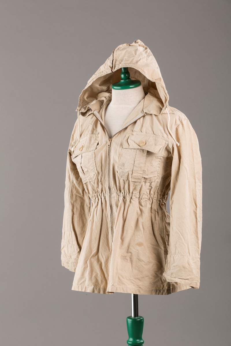 Form: Vanlig anorakkfasong med strikk i livet. 2 brystlommer med knapp.
Hette med glidelås på topp midt, snøring.
Glidelås helt ned foran.
Innermansjett med strikk i ermene.