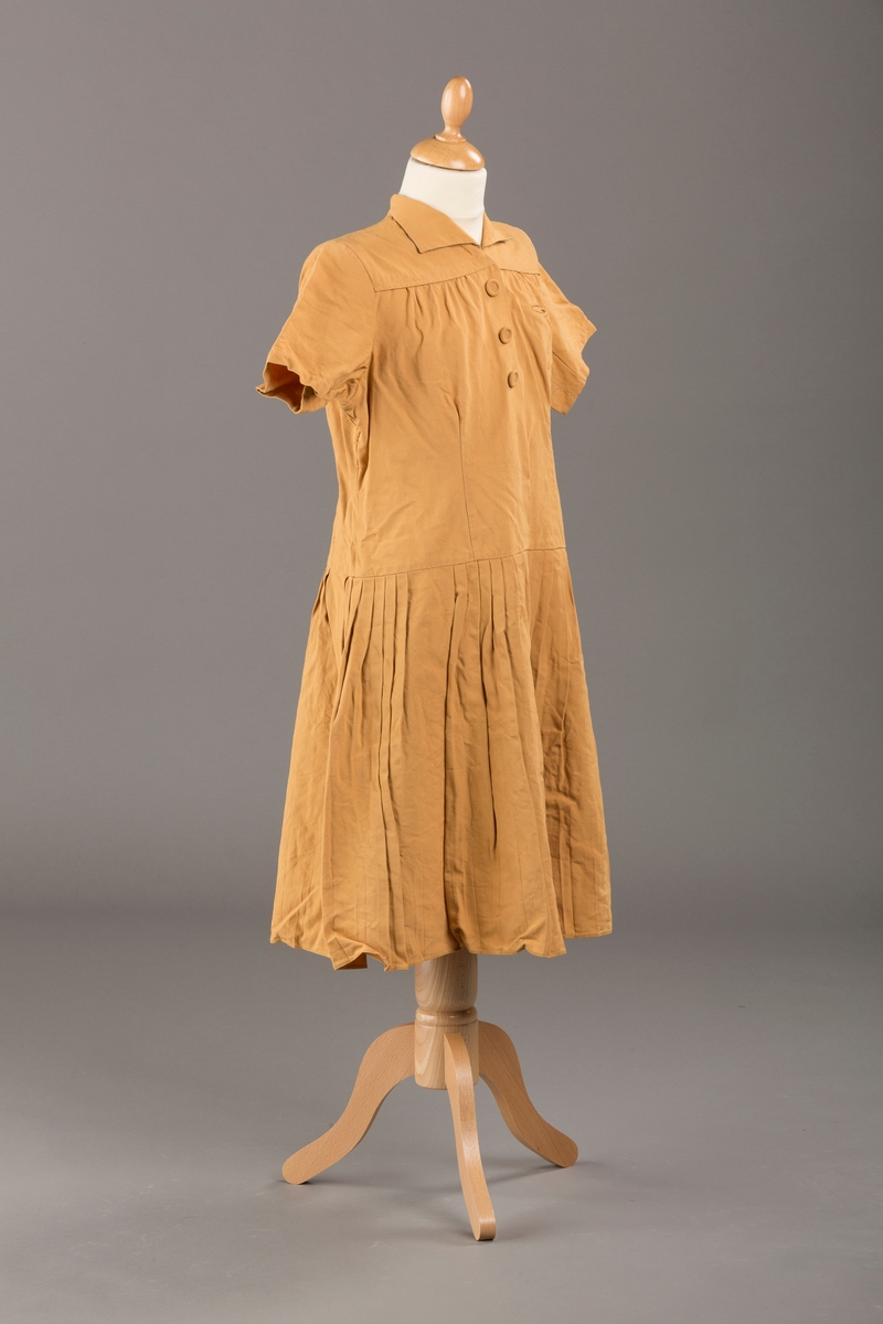 Form: Korte ermer, livet rynket til bærestykke foran, plissert skjørt, tre pynteknapper, en trykknapp, forlomme.