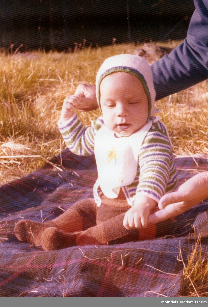 Filten ligger på en äng med halvhögt gräs år 1974. Man ser händerna av en vuxen person som håller i Martin (född 1974-01-12) som stöd. Babyn är givarens storebror.