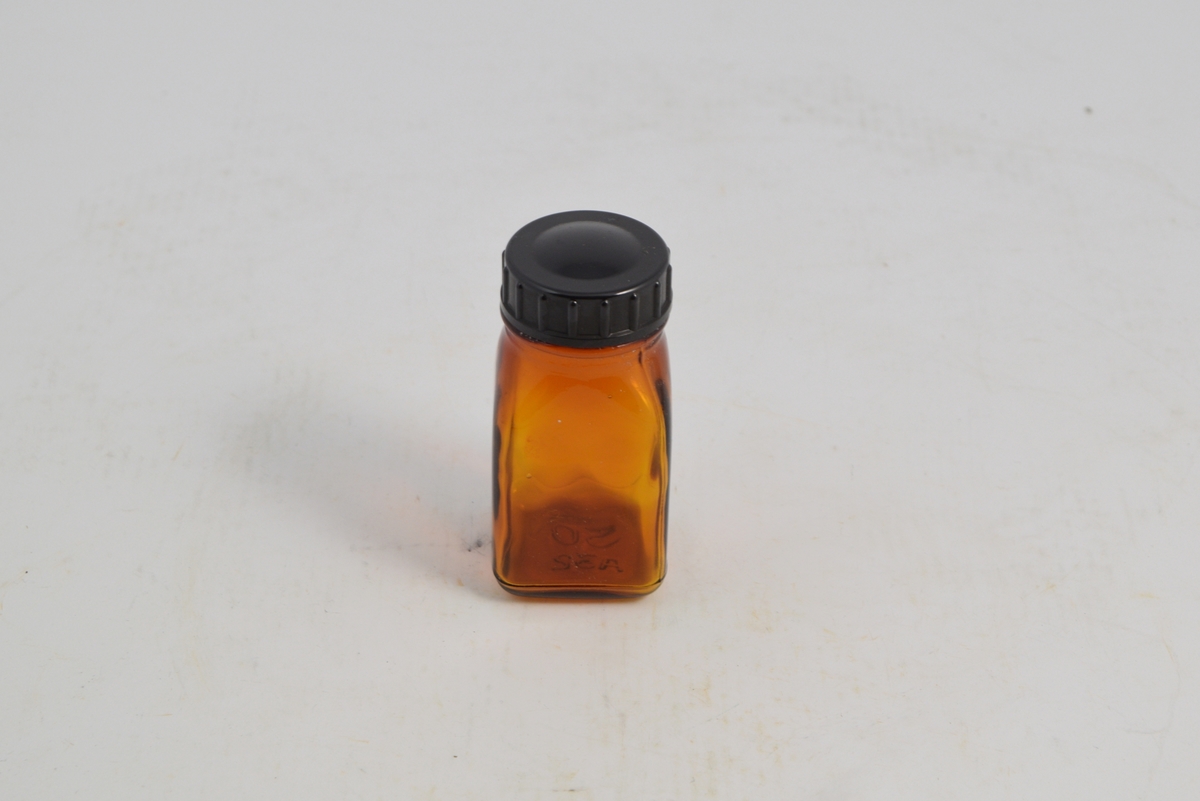 Brun transparent firkantet tablettglass med sort rundt bakelittlokk, skru. Brukt til oppbevaring og salg av tabletter, ca. 1950-1960.