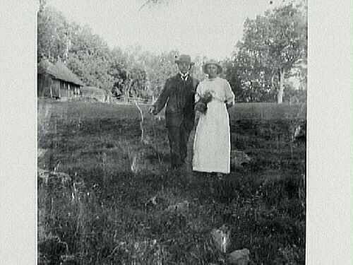 Ungt par (kvinna utklädd till man?) står i en gräsbacke utanför en trädomgärdad gård. Kvinnan håller i en bukett med ormbunkar.