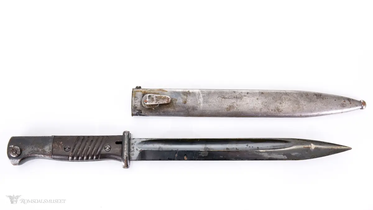 Metallhåndtak med lang pommel og geværfeste med fjæret knapp i bakkant. Serratert tregrep som er festet med 2 skruer. Langt spisst stålblad med egg og blodrand. Metallslire med kule enden og krok for festing til grunnutrustning/belte.