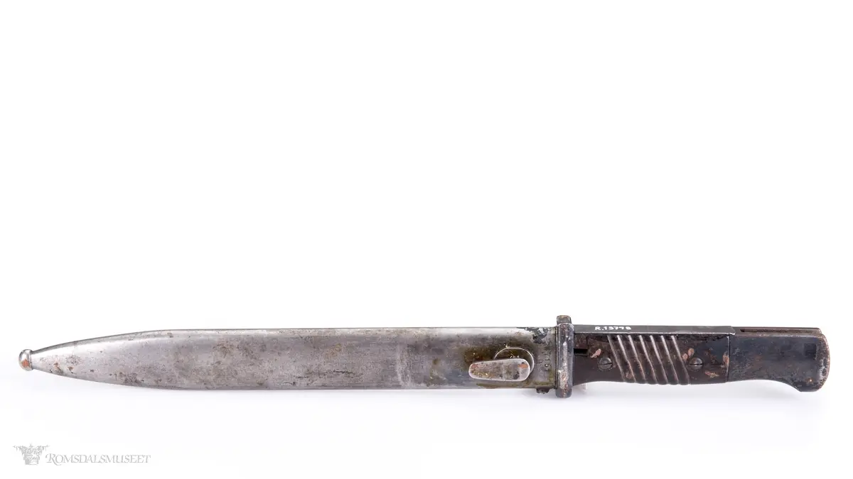 Metallhåndtak med lang pommel og geværfeste med fjæret knapp i bakkant. Serratert tregrep som er festet med 2 skruer. Langt spisst stålblad med egg og blodrand. Metallslire med kule enden og krok for festing til grunnutrustning/belte.