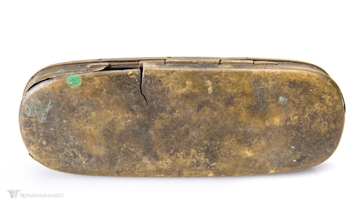 Rektangulær messingeske med avrundede ender. Har en påsatt og påklinket utenpåliggende bunn og utenpåliggende, hengslet lokk