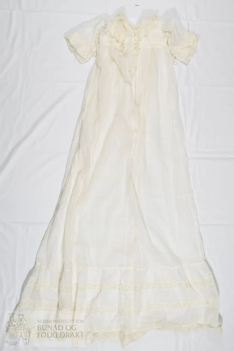 Lang dåpskjole i hvit bomullslerret med påsydde kjøpeblonder. Livstykket er sydd av to lag med tynn bomullslerret. Sydd til skjørtet med en bred linning. Ermer sydd rett i, med rynker langs hele ermeåpningen. 

Åpning midt bak med splitt et stykke ned på skjørtet, knyting øverst med bånd. Forstykke og de to bakstykkene er sydd sammen på skuldrene. Halslinning er brettet ned dobbelt på baksiden, og er sydd ned på maskin. Bred blonde i halsringingen som er sydd på med maskin. Blonden er maskinlaget, og er av bomull. Den er noe rynket til halslinningen. 

Ermet er sydd av et enkelt lag med tynn bomullslerret, og har påsydd en løpegang for knytesnorer på vrangen. På vrangen er også festet en bred blonde som stikker ut av ermeåpningen.

Skjørtet er tett rynket til linningen i livet og sydd ned med maskin. Skjørtet er sydd av fem høyder, sydd sammen med maskin. Et felt midt foran er dekorert med blonder langs skjørtehøydene på hver side, og som går hele veien ned til skjørtekanten. I tillegg er horisontale blondebånd sydd inn mellom felt av bomullstoffet i skjørtehøyden midt foran. Alt er sydd på maskin. Nederst på skjørtet er det påsydd en bred kappe, som er lett rynket til skjørtet. Kappen har mellomverk av maskinblonde.