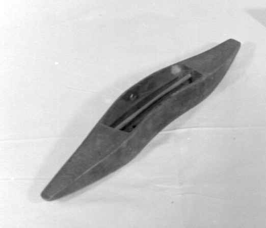 Avlang og buet vevskyttel av tre med uthuling på midten og trådspole av tre. På hver side er det skyttelen dekorert med hjerter. Den ene siden har inskripsjon MAD og den andre siden årstallet 1823