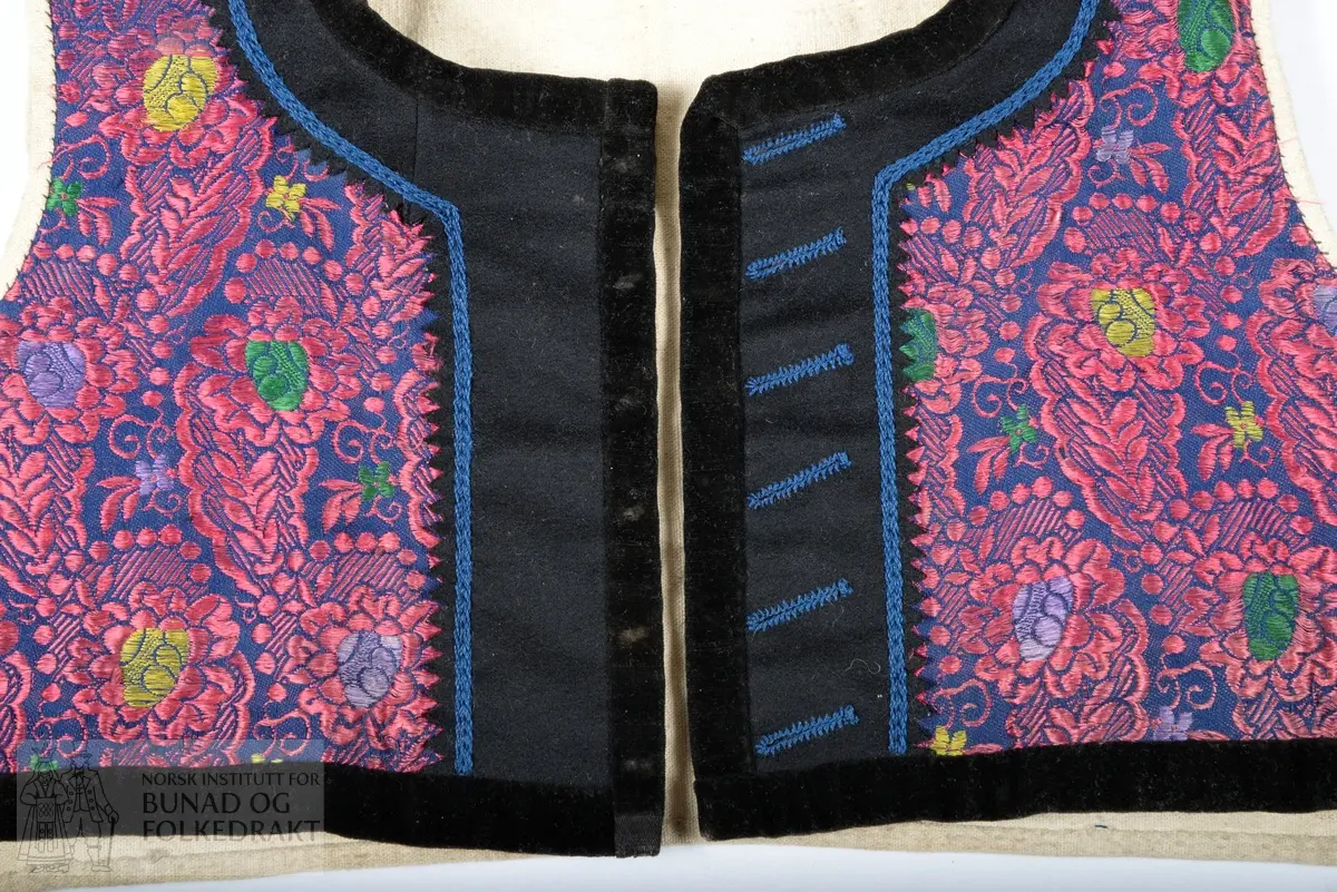 Vest, sauma av silkedroguet. Blå satengbotn med rosa flotterande trådar og innvove små blomster i ulike fargar. Framkant, krage og nedkant er påsydd ein smal svart kant av bomullsfløyel. Innanfor denne i framkant og krage er det påsydd eit breitt stykkje svart tynn fast klede. Denne er klipt med taggar etter sårkanten, pryda med tre tette konturstingsrader med blå ulltråd. 
Kragen er fora med tjukk brunsvart vadmål, sydd med to langsgåande avstivande stikningar. Ryggen er ufora, sauma av litt grov bomullslerret. 
Same type for i framstykkja. 
I ermholet på framstykkjet er foret bretta dobbelt rundt på framsida, og sydd ned med små javne faldesting. Sidesaumane er drage eit godt stykkje inn på kvart framstykkje. 
Eine framkanten har påsydd 6 stk skråstilte knapphol sauma av same type blå ulltråd. Merkjer etter knappefeste gjennom fløyelskanten på andre framstykkjet. 
I sidesaumen er hovudstoff framstykkje og ryggfor sydd saman med attersting, deretter er for framstykkjet bretta inn og sydd ned 1 cm bak den fyrste saumen, med faldesting som syner som prikk på retta.