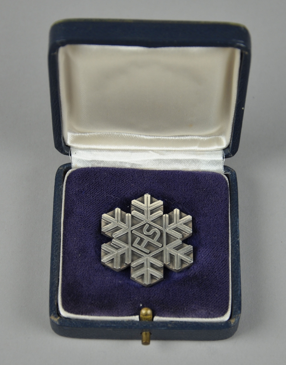 Medalje (nål) fra VM i Squaw Valley 1960. Medaljen er formet og mønstret som snøfnugg. 
Medaljen ligger i et blått etui merket FIS