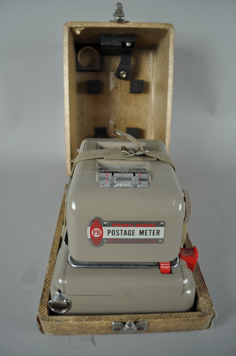 Frankeringsmaskin av brunt metall. 

Maskinen er plassert i trekasse med fiskebensmønstret utside. Kassen har håndtak.