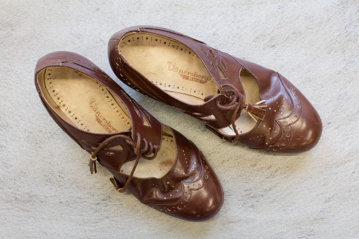Ett par damskor i brunt läder. Skorna har läderklack. Bred slejf med snörning fram med genombrutet mönster. Perforerad infälld bit vid hälen och vid tån. Snörningen är försedd med tofsar.
På undersidan av skon vid valvet står 5HA.
På sulan i skon står Vänersborgs sko.