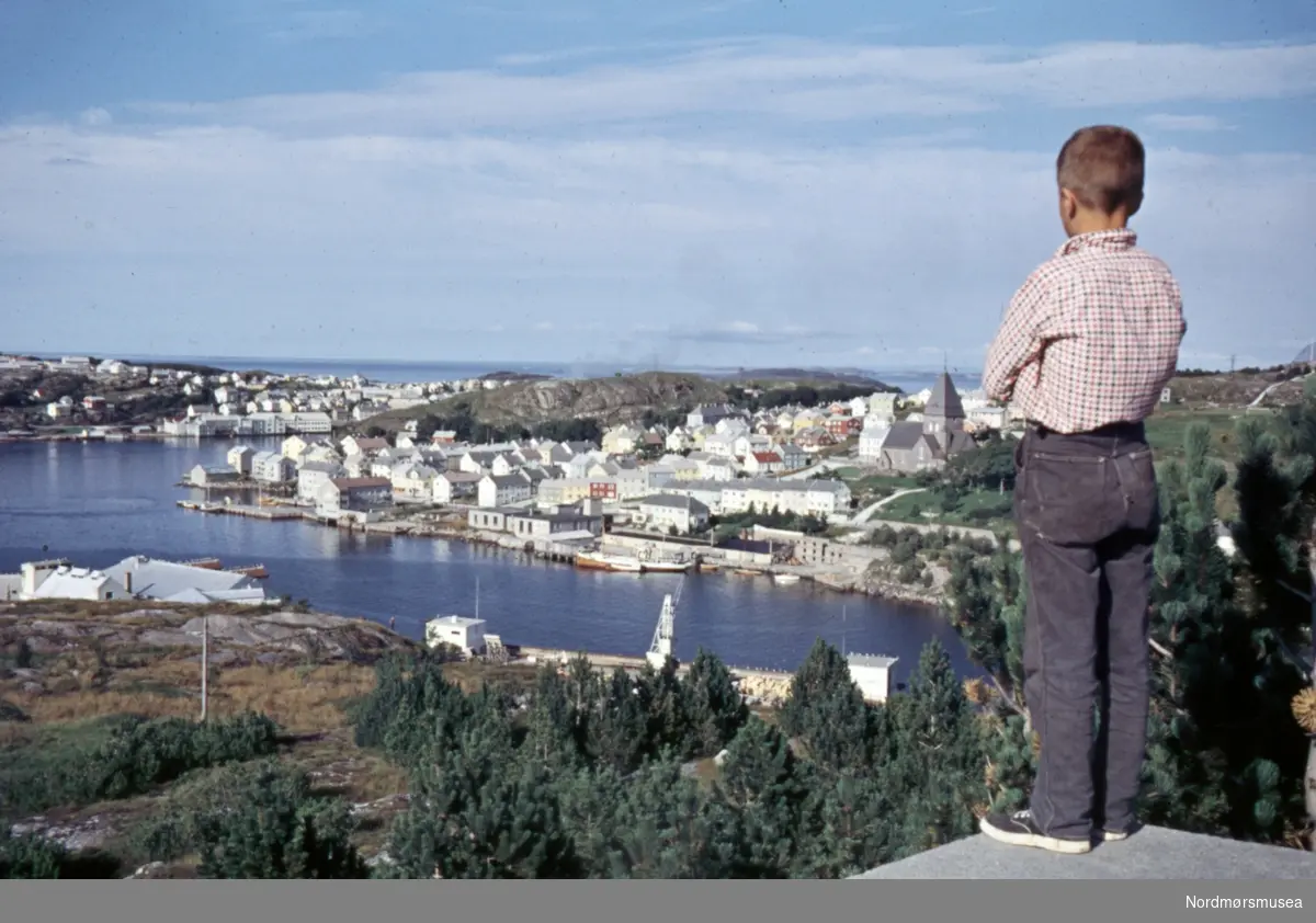 Foto av en gutt som står å ser i retning Nordlandet i Kristiansund. Bildet er fotografert fra Innlandet. Fotograf er Kjell Haug og er den som står bak samlingen med fargefoto/dias fra Kristiansund og Grip. Bildene dateres til omkring 1950-1970. Fra Nordmøre museums fotosamlinger.