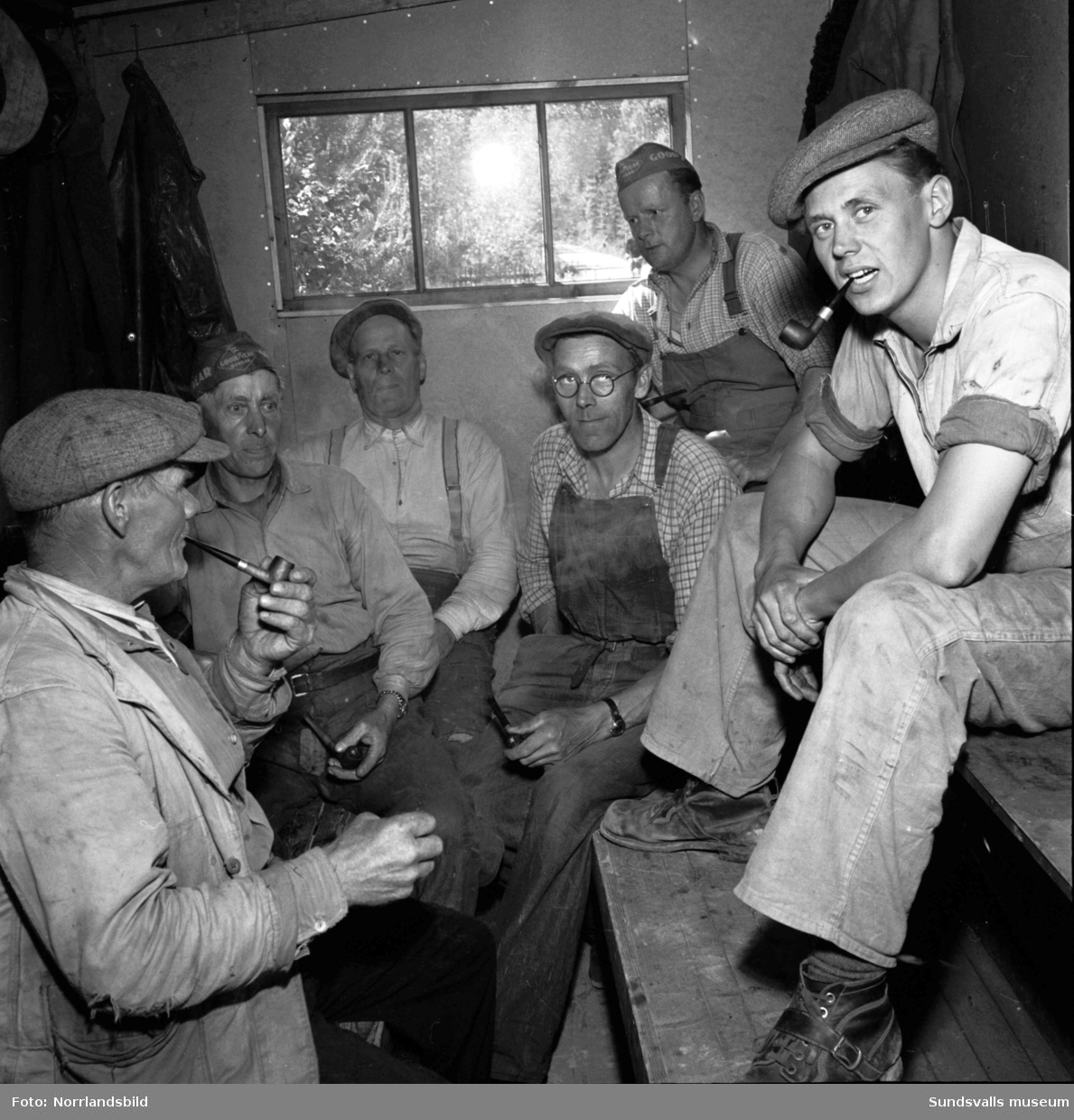 Vägarbetare vid bygget av vägen till Bogrundet i Timrå. Sex piprökande män som tar en paus i en raststuga, barack.