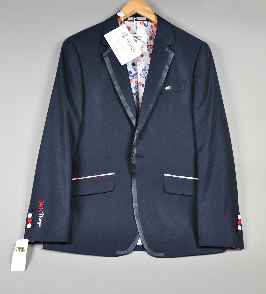 Mørkeblå dressjakke av ull/silke, foret med samme silkestoff som i damekjolen og tørkleet, et
hvitt silkestoff med "rosemalt" mønster.Jakken har fire innerlommer, en med knappelukking.
Utvendig to jakkelommer med klaff foret med silkestoffet, en brystlomme med påbrodert
"traktorlogo", Mørkeblått kantebånd fra kraven og ned. Lukket med en stor stoffknapp på ermene, fire ganger to mindre stoffknapper i hvitt (2), rødt og blått. På høyre erme brodert:Heia Norge i henholdsvis rødt og hvitt.
Str.:48