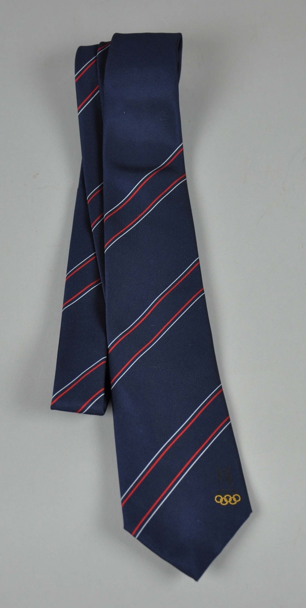 Blått slips  med NOK/NIFsin  logo innvevd i sort,  de olympiske ringene innvevd  i lys  sennepsgult,plassert til høyre nederst på slipset.Diagonale striper i hvitt og rødt.To like ,som forrige nr.
