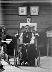 Eldre kvinne fra Otta i damaskstakk sittende i rullestol. Gu