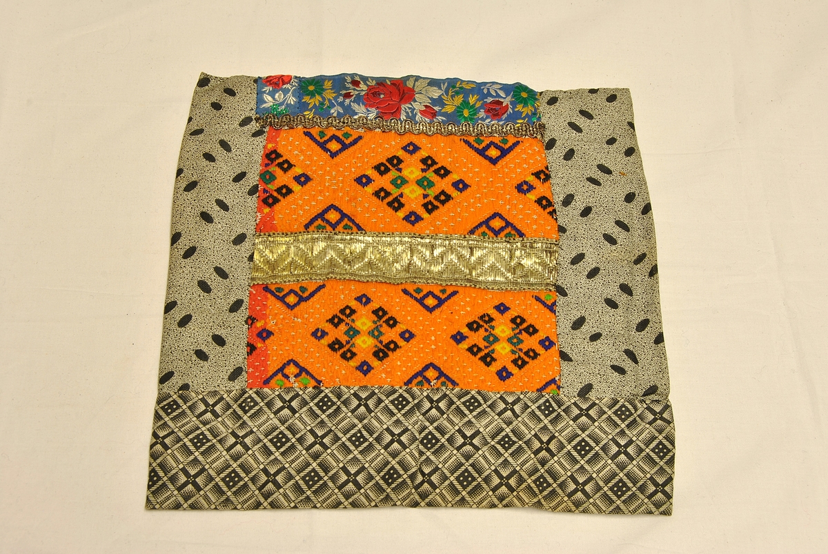 Nær kvadratisk form. Sydd sammen av flere tøystykker. Broderi i front. Pyntebånd oppe og i midten av broderiet.