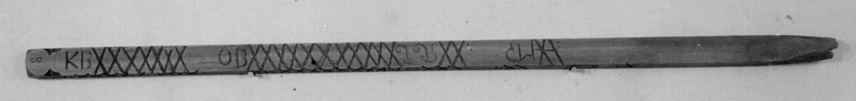 Målestokk av tre som er spiss i den ene enden. Det er inngravert mange kryss, streker og punkter på alle fire langsider med signaturer for hver rekke av merker.