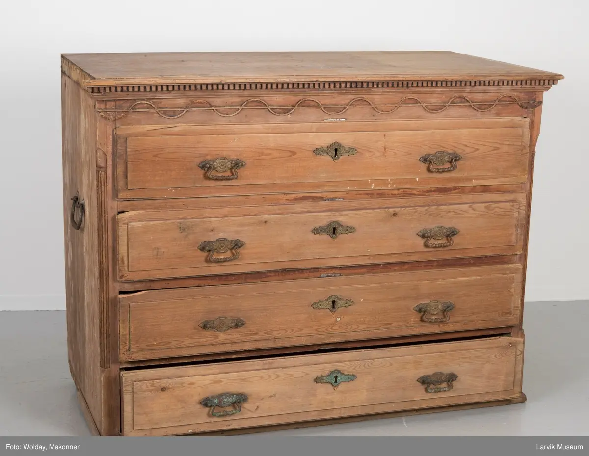 Kommoden har 4 skuffer. 2 beslag for å dra ut, 1 beslag for nøkkelhull, messing. Utskåret dekor under øvre pyntelist "tanngard" rundt på 3 kanter. Øvre skuff utskåret med bølgebånd med rett linje som bryter bølgene. Utskjæring kun i front. To av kommodens hjørner har utskåret søyle. Håndtak på kortsidene for å bære i.