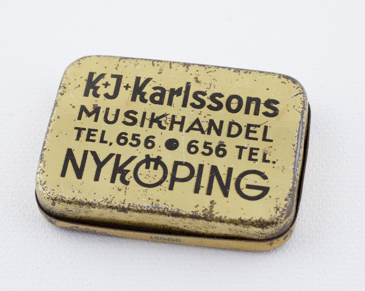 Ask av plåt.
Fyrkantig modell med avrundade hörn och dekor i blått och guld. Förvaringsask för grammofonnålar. 
Text på locket, "K.J. Karlssons Musikhandel Nyköping".