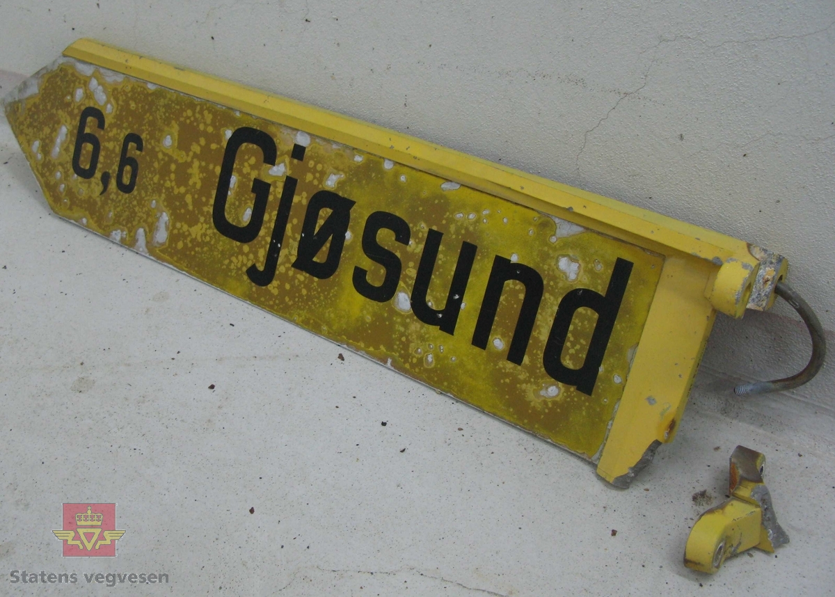 Vegviserskilt. Skiltplata er lysreflekterende gul med svart tekst "Gjøsund 6,6". Selve skiltplata er lagd av en aluminiumsplate der festet og kanten øverst er støpt i en del. Skiltet har vært festet til 3 toms stolpe ved hjelp av to bøyler. En bøyle henger på skiltet
