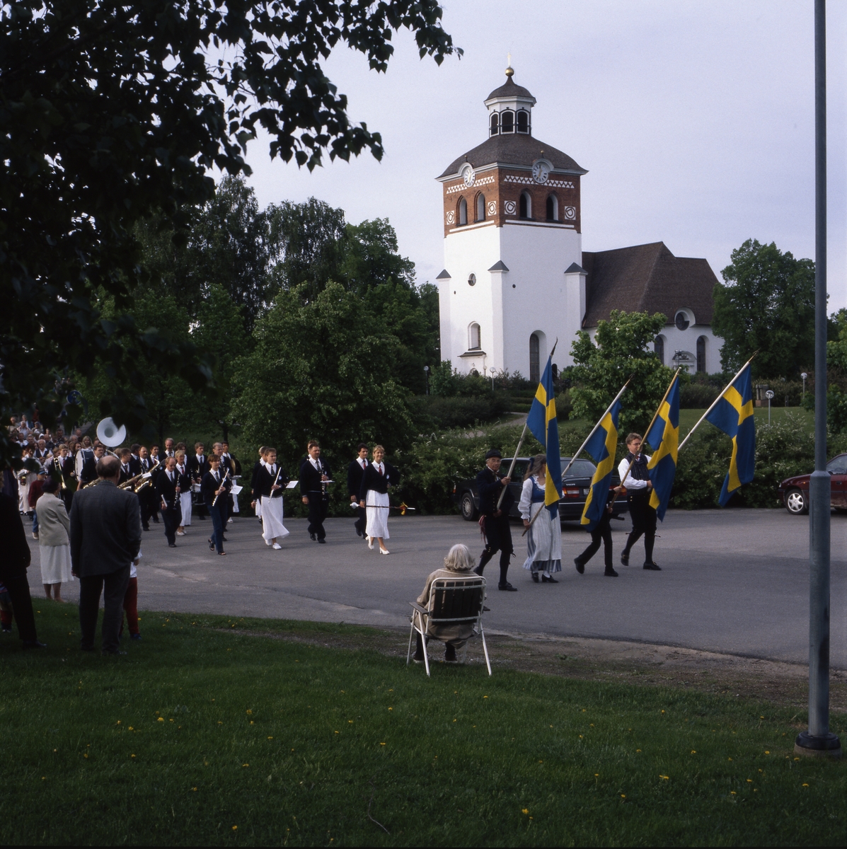 Nationaldagen i Bollnäs 6 juni 1999. Fanbärare och musikanter tågar förbi kyrkan. En äldre kvinna sitter i en trädgårdsstol bredvid vägen och ser på.