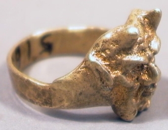 Fingerring av mässing. Ringen har ett utvidgat framstycke utsmyckat med ett mefistofelisansikte i relief.