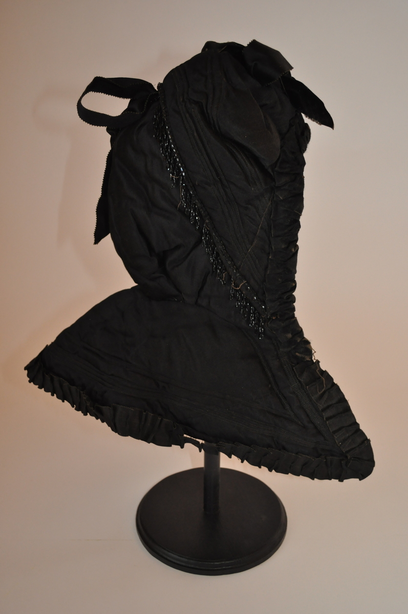 Kyse, "kråke" av svart silke med dekor av svarte silkebånd og perlefrynser. Kysa lukkes med hekter og snøring foran.