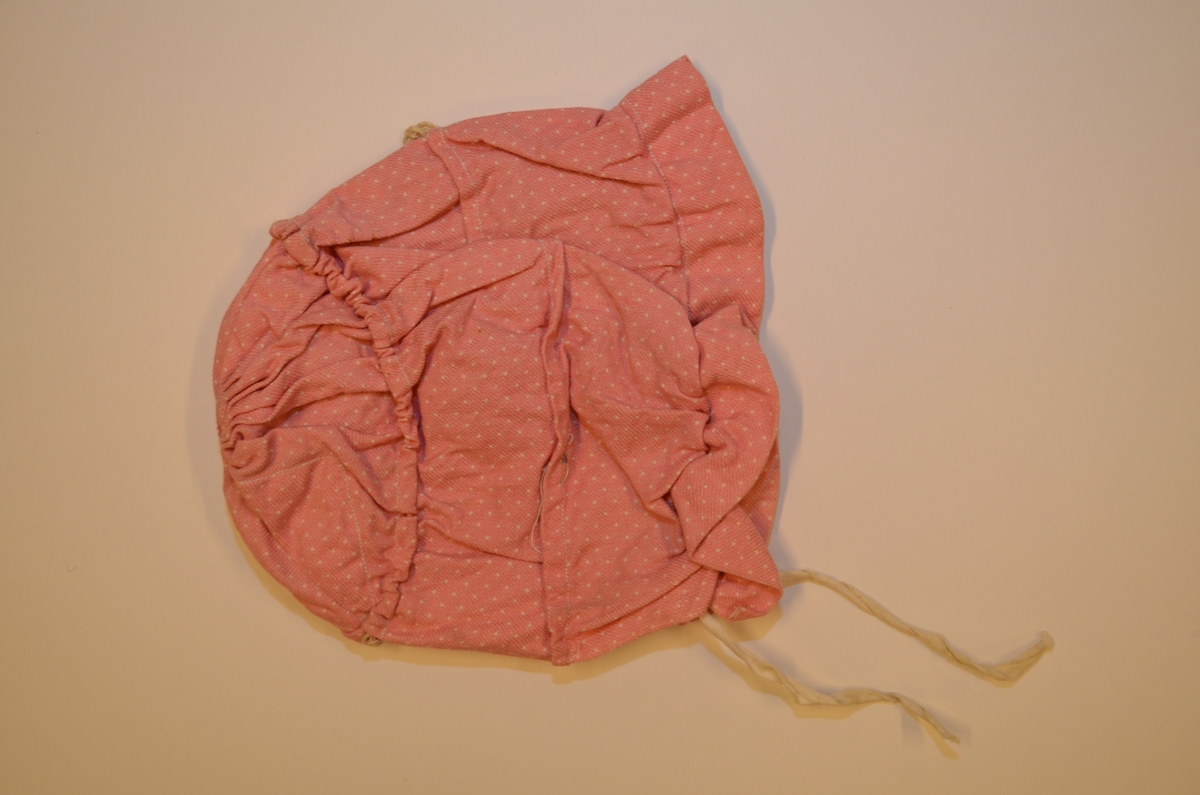 Barnelue i rosa kypertvove bomullstøy med kvite prikkar. Eittstykkslue med tre påsydde løpegangar som er rynka i saman. Foldelagt i "pullen", kappe i framkant. To knyteband. Maskinsydd.