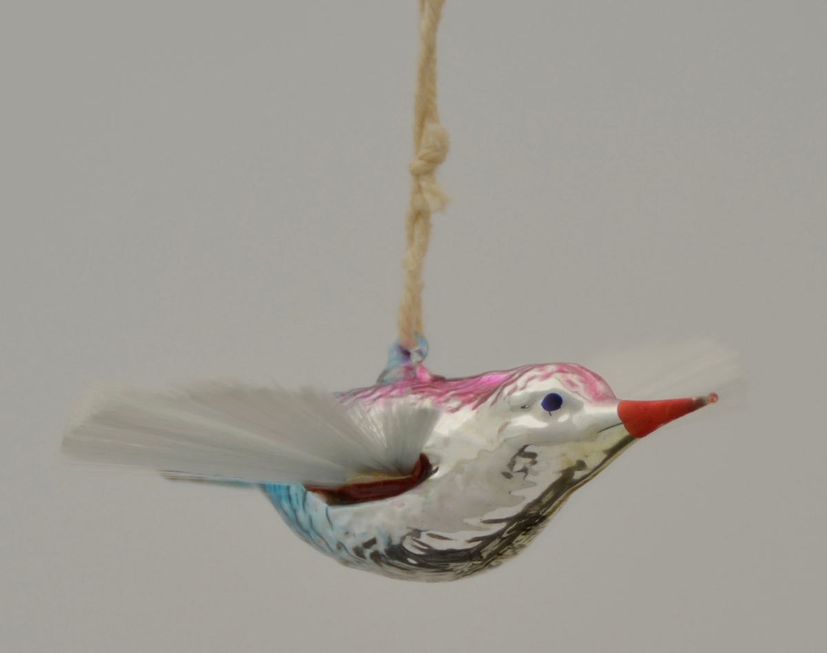 Juletrepynt. Glasfugl med vinger som ei vifte av silkeaktig, stiv tråd. Glaset er sølvfarga i botn med lilla og blått på kroppen og raudt nebb. Tråd gjennom oppheng på ryggen.