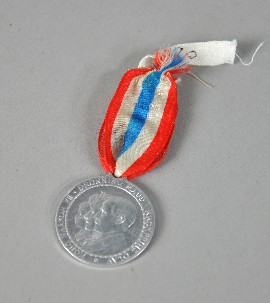 Medalje av aluminium med sløyfe i rødt, hvitt og blått. Medaljen viser portrettene av kong Håkon, dronning Maud og kronprins Olav, med tekst rundt kanten.