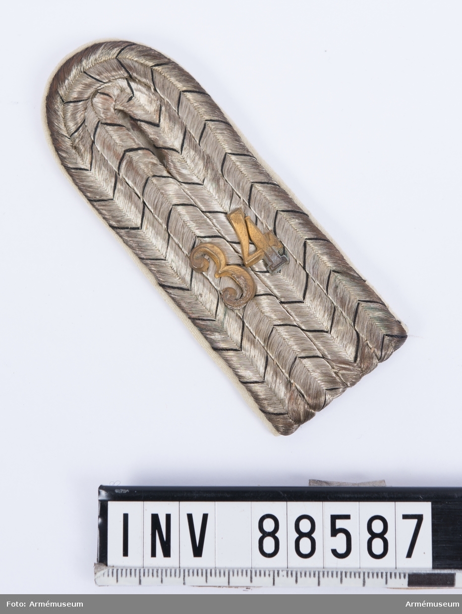 En axelklaff med fyra vävda snören av silvertråd med svarta inslag (preussens färger) och nummer "34" i mässing. Kantningen är av vitt kläde. Axelklaffen användes till den fredstida uniformen.