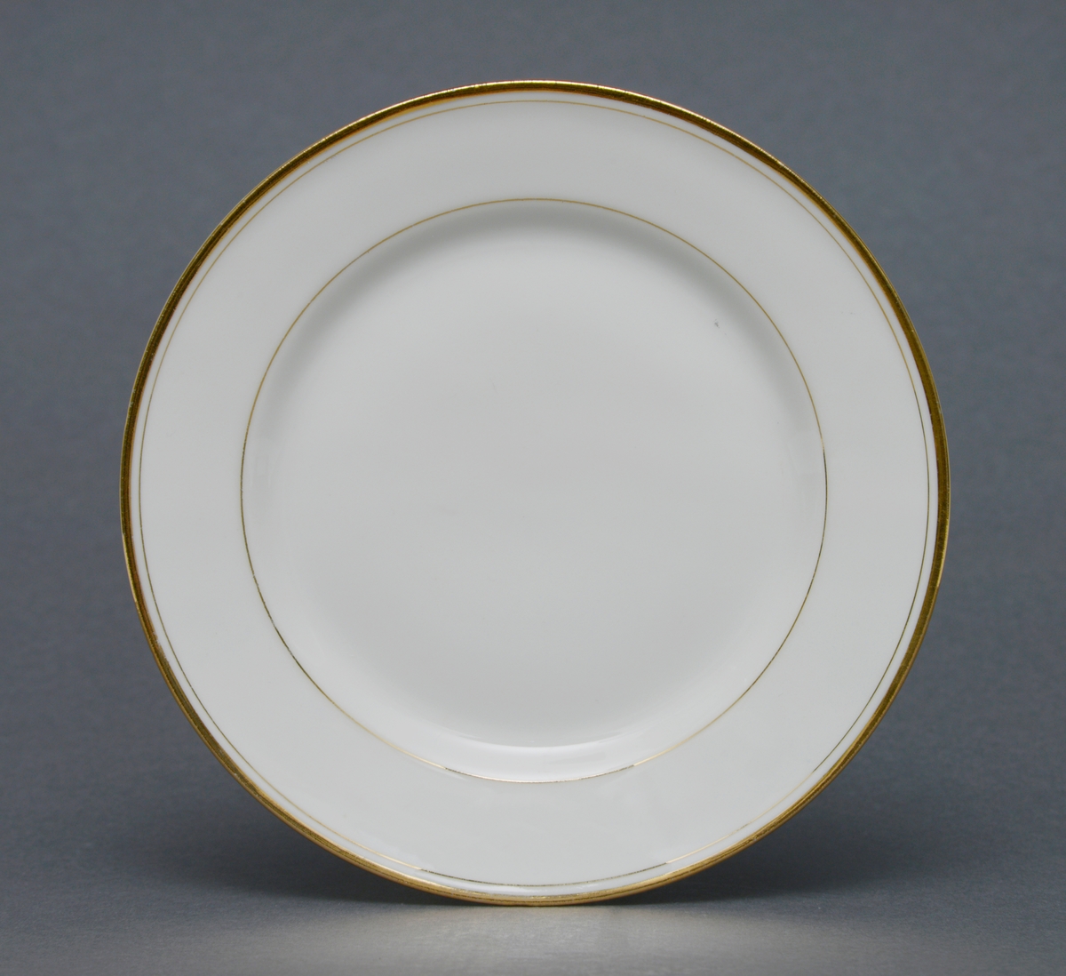 Sirkulær tallerken i porselen med glasur. Dekorert med èn gullforgylt ring rundt kant, og èn mot midten av tallerkenen ved fordypning.