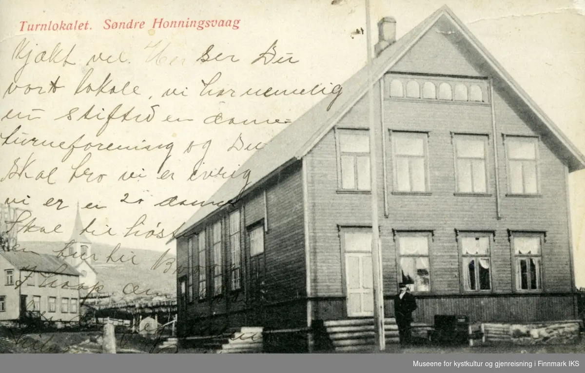 Postkort. Turnlokale i Honningsvåg. I bakgrunnen kirka og fiskehjeller med tørrfisk. Omkring 1914.