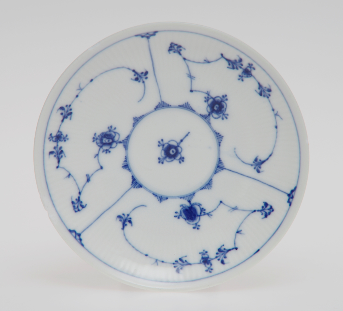 Sirkulært tefat i porselen med glasur. Rifler i porselenet og dekorert med stråmønster i blått.