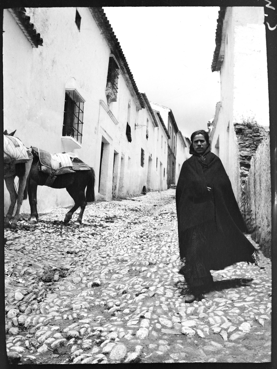 Kvinna på kullerstenslagd gata. Två åsnor i bakgrunden. Troligen Ronda, Spanien.