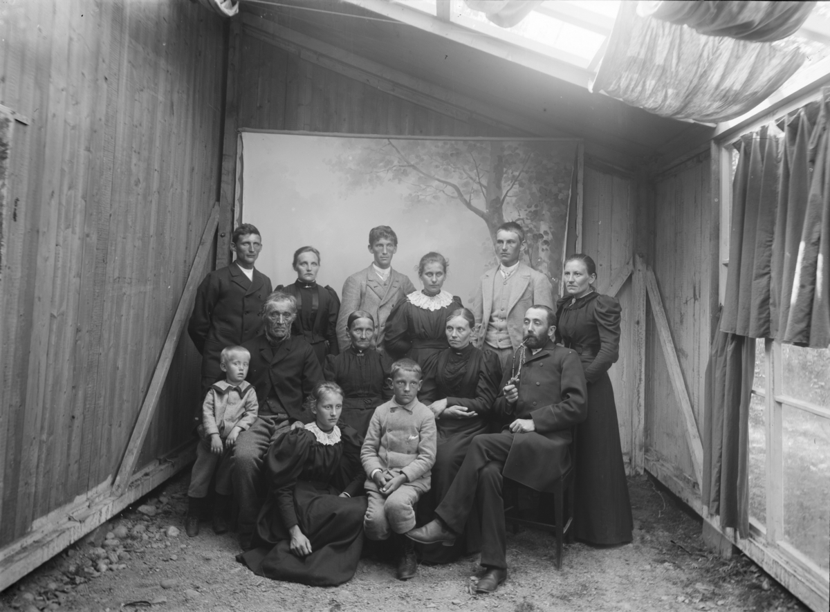 Familiebilde med 13 personer og tre generasjoner. Atelierfoto (provisorisk ved reisverksbygning) fra Gudbrandsdalene eller sidedalene i tidsrommet 1890-97