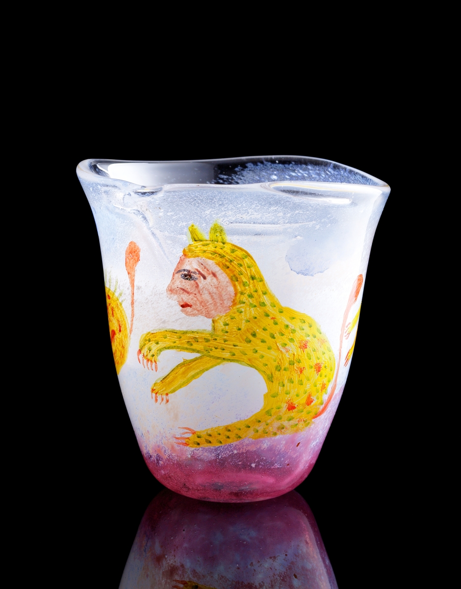 Vas i en unikatserie. Detta exemplar nr 110.
Formgiven av Ulrica Hydman-Vallien.
Tillverkad under första halvan av 1970-talet.
Utställd i "Glas 75".
Svagt utvidgande vas, något oregelbunden form. Treuddig, oregelbunden kant.
Vasen är till stora delar täckt av en vit pulverfärg. Mot botten en rosa pulverfärg. 
Målad dekor i form av tre gula kattdjur med grönprickig päls och människoansikten.