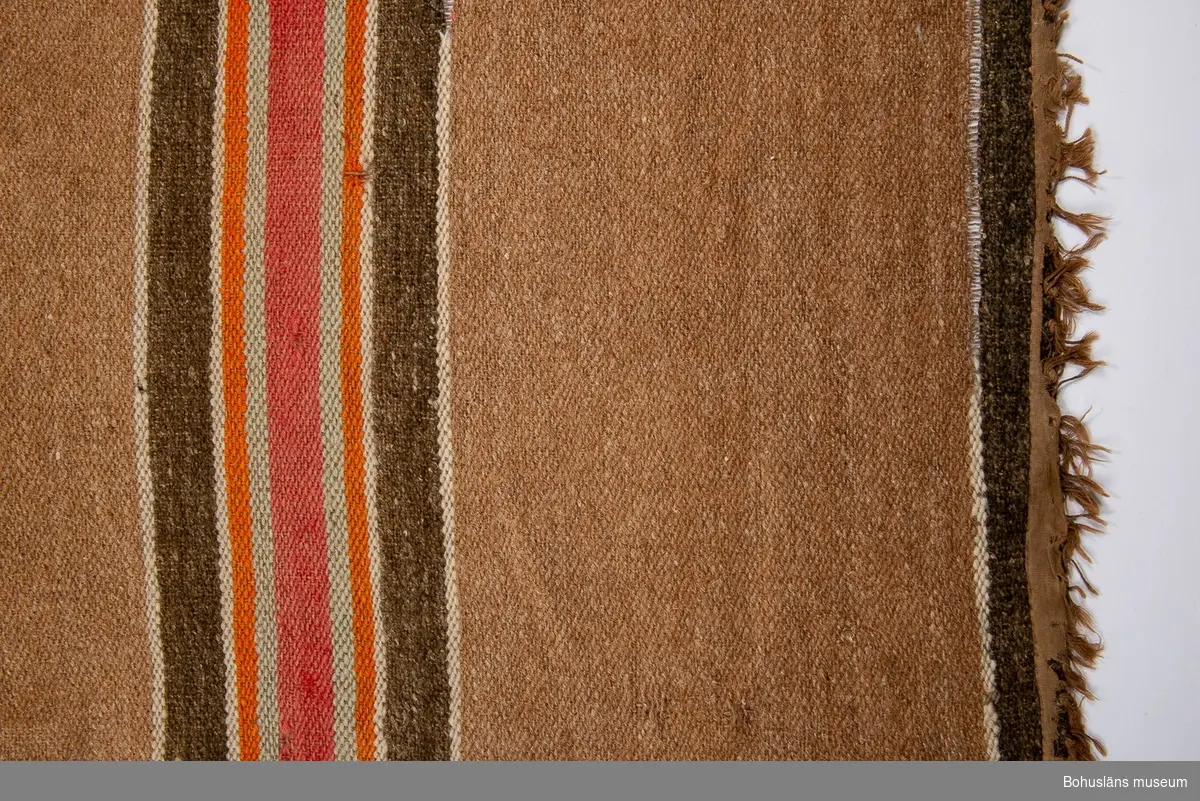 Beige-brun. Tvärrandig i rött, ljusblått, orange, vitt och grått.
Denna matta är en av fyra stycken som låg i en fyrkant i salen hos givarens mormors mor på Orust från ca 1890 och framåt tiden.
Se bilaga i Bilagepärmen.