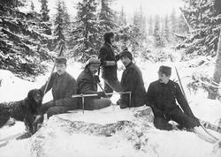 Tømmertaksering i fjellskog.  Fire menn sitter i snøen bak e
