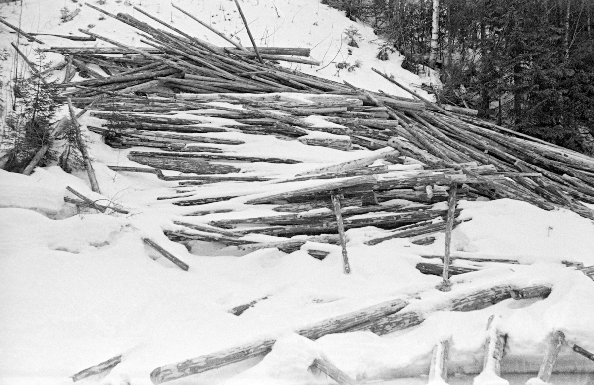 Vinteropptak av tømmervelte i Hovda-vassdraget i Åmot i Østerdalen.  Fotografiet er tatt på tvers av et is- og snødekt elveleie mot en forholdsvis bratt elveskråning, der barket tømmer øyensynlig er rullet ned mot elveleiet i løpet av vinteren.  Stokkene ligger noe hulter til bulter, men i hovedsak parallelt med vassdragets strømretning.  Bakenfor velta, øverst til høyre i bildeflata skimtes forholdsvis tett granskog.  Bildet ble tatt i april 1952.  Dette var en måte å levere tømmer på som Glomma fellesfløtingsforening ønsket å komme bort fra på dette tidspunktet, jfr. SJF. 1990-00300. Hovda og dens sideløp har et samlet nedslagsfelt på drøyt 190 kvadratkilometer.  Elva er fløtbar fra innsjøen Storhovden og nedover, men tømmer som ble levert på den øvre delen av denne strekningen nådde ikke alltid fram til Glomma i løpet av en sesong.  Først fra sammenløpet med sideelva Hemla og østover ble fløtinga i Hovda ansett for å være «årssikker».
