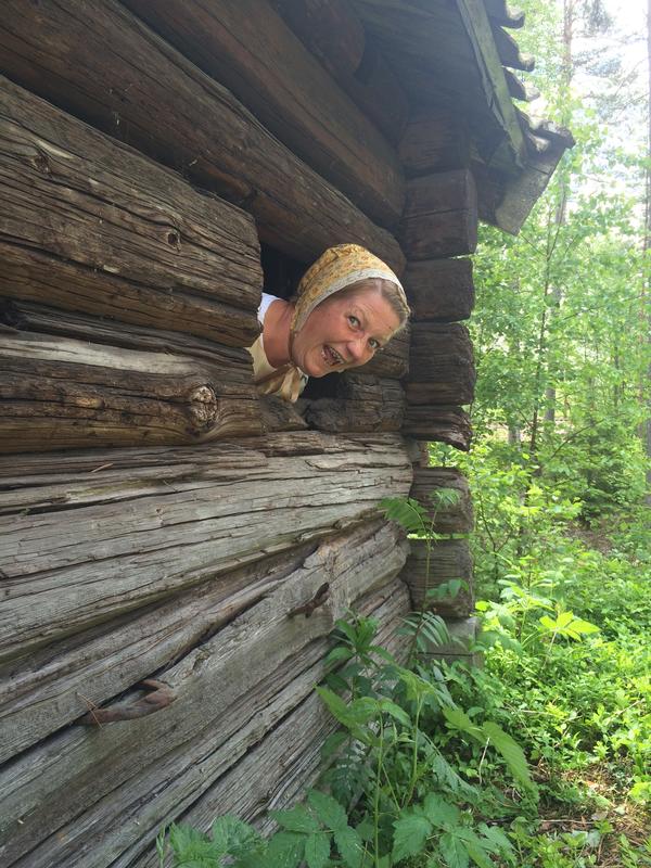 Mette Skogbakken titter ut av et vindu i et tømmerhus. Hun smiler og har på en gul kyse.