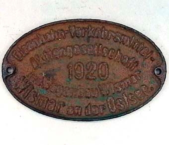 Oval skylt av järn med gröna färgrester. Text: "Eisenbahn-Verkehrsmittel-Aktiengesellschaft Wagenbau Wismar, Wismar an der Ostsee 1920".