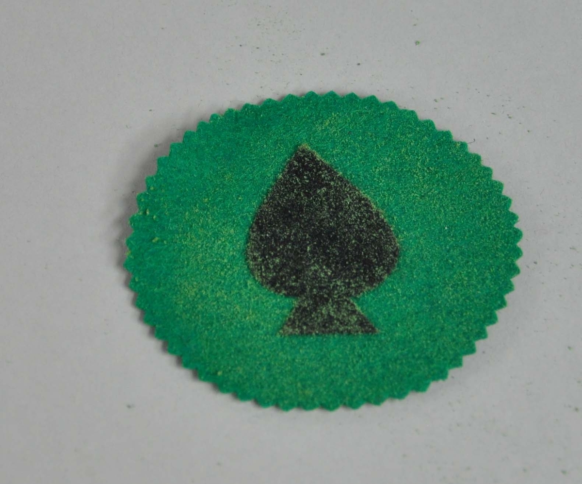 Rund brikke av grønt stoff, med bølgete kant. På midten er symbolet for sparkortet.