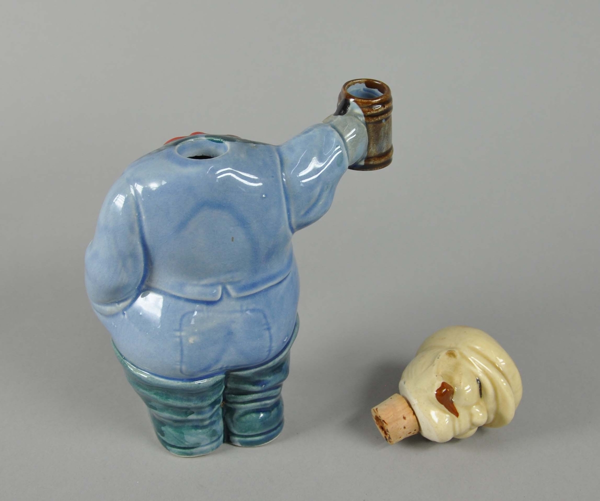Karaffel av glassert keramikk. Forestiller mann med ølkrus i hånden. Hodet på mannen fungerer som lokk til flasken, det er satt inn en kork under hodet hvor halsen er.