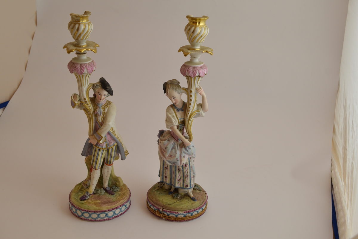 To lysestaker i form av statuetter av en mann og en kvinne i typisk 1700-talls fransk overklasse bekledning, De holder begge hver sin blomst som fungerer som stearinlysholdere.