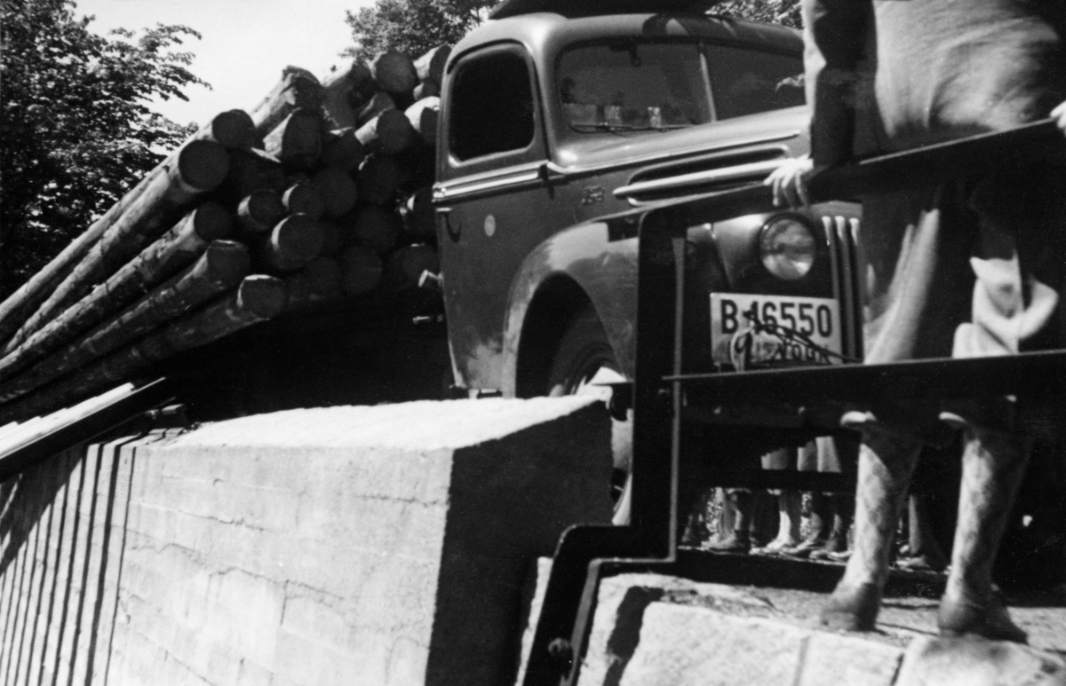 Tipping av lastebiltransportert tømmer i Haldenvassdraget.  Fotografiet under Norsk forstmannsforenings utferd i denne regionen i juni 1949.  Det er tatt fra vassdragssida mot en betongstøpt mur som rager en drøy halvmeter over en bakenforliggende velteplass, der vi skimter førerhuset på en lastebil med registreringsnummer B 16550.  Dette var en Ford av årsmodell 1946-47.  Vi ser også hvordan tømmerlasset, tilsynelatende i buntet form, er i ferd med å veltes over på en del skinner som var lagt på skrå fra overkanten av den nevnte betongmuren mot det nedenforliggende vassdraget (jfr. SJF-F. 006110).  I forgrunnen til høyre ser vi beina og ryggen til en mann som var kledd i nikkersbukser og dressjakke.  Etter 2. verdenskrig ble bruk av lastebiler i tømmertransporten stadig vanligere.  Ved de store fløtingsvassdragene ble det opparbeidet velteplasser, der tømmer som var hentet langs de mange nye skogsbilvegene og kjørt til hovedvassdragene og slått uti, slik fotografiet viser.  Haldenvassdragets fellesfløtningsforening later til å ha vært tidlig ute med slike anlegg.