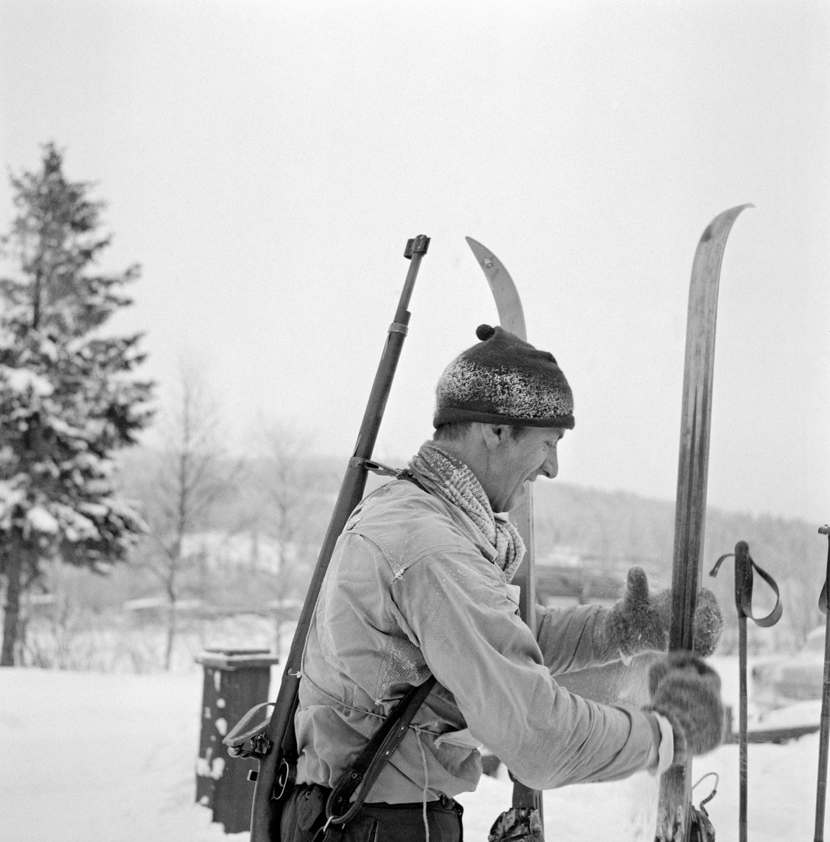 Fra det første offisielle kretsmesterskapet i skiskyting, som ble arrangert på Rasta ved Kongsvinger 8. januar 1961.  Rennet gikk i bitende vinterkulde, det skal ha vært 29 kuldegrader da de 22 påmeldte løperne ble oppropt før start. Løypa var 8-9 kilometer lang og hadde fire standplasser, der det ble skutt på ulike hold (250, 150, 200 og 100 meter).  På dette fotografiet ser vi Arvid Nyberg (1928-2022) fra Trysil sette fra seg ski og staver. Han var kledd i en lys anorakk.  På hodet hadde han ei rimete topplue og hendene var bare i kulda, rundt halsen et skjerf og på hendene et par digre ullvotter. Trysil-løperen fikk nest beste langrennstid i klasse A, men endte på sjetteplass fordi han bare traff 7 av 20 blinker i dette rennet. Nyberg var lagkamerat med toppløperen Hallgeir Brenden (1929-2007), og var med på å vinne tre stafettgull i langrenn for Vestre Trysil idrettsforening.  Gjennom idretten kom han også inn i organisasjonslivet, og derfra kom han med i lokalpolitikken.  Arbeiderpartimannen Arvid Nyberg var ordfører i Trysil fra 1972 til 1999, altså i 27 år.  Han har seinere hatt et organisatorisk engasjement i Hedmark Jeger- og Fiskerforening.