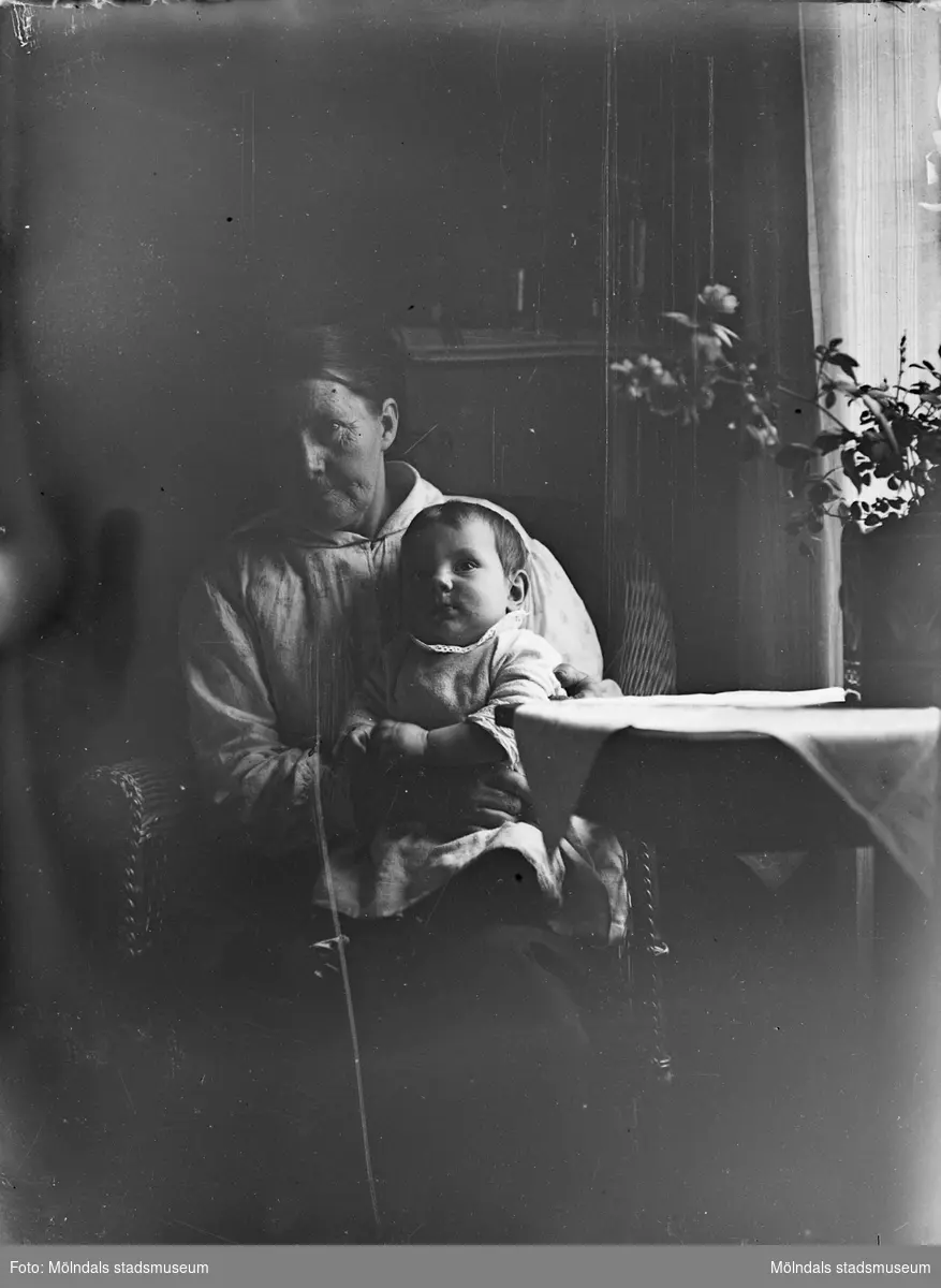 I hemmiljö där en äldre kvinna sitter med ett litet barn i knäet, troligtvis 1920-30-tal.