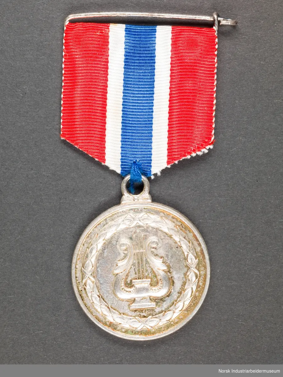 Rund, sølvfarget medalje festet i tekstilbånd i Norges nasjonalfarger. Nål festet til tekstilbånd. Medaljens motiv er på den ene siden en harpe omgitt av en bladkrans og på den andre siden tekst.